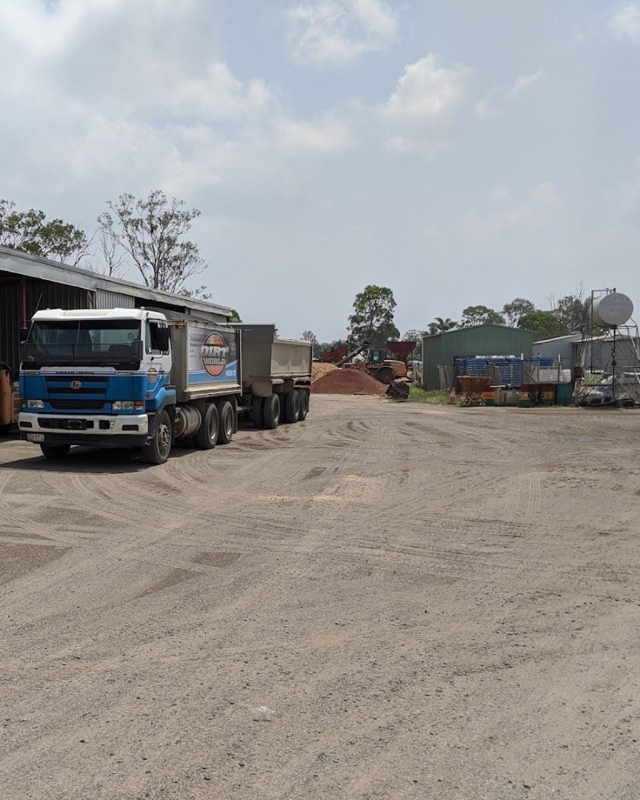 A tipper truck and trailer parked at the yard beside the sheds, with stockpiles and a loader working behind
